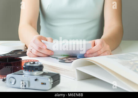 Married or engaged girl in dress sits at white desk with film camera and film strip and puts printed photographs into family album Stock Photo