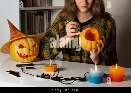 Young female person sitting at working table in living room makes drawing with marker on pumpkin to make Jack O' Lantern Stock Photo