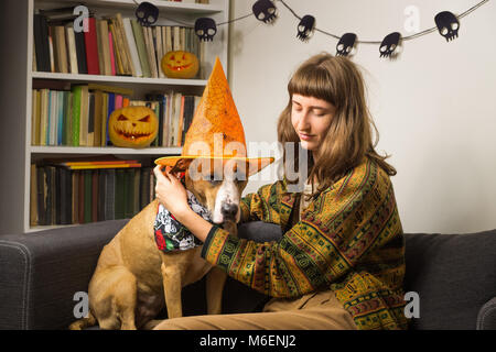 Girl and her pet sit on sofa in living room with jack'o'lantern pumpkins in background, dog wearing halloween costume hat and skulls bandana Stock Photo