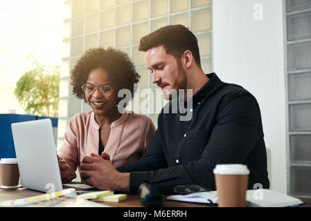 Two smiling businesspeople sitting together at a table in a modern office talking and using a laptop Stock Photo