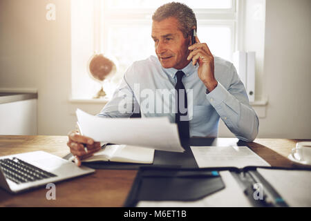 Smiling mature consultant discussing documents over the phone while working at his desk in an office Stock Photo