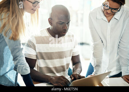 Three diverse design colleagues working online together on a laptop at a desk in a bright modern office Stock Photo
