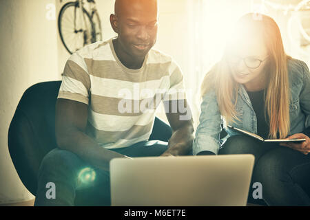 Two diverse colleagues smiling while sitting together working on a laptop in a stylish modern office Stock Photo