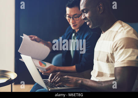Two diverse work colleagues sitting together on an office sofa discussing documents and working on a laptop Stock Photo
