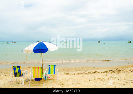 Isolated beach with three colorful beach chairs and a blue and white umbrella. A small wave breaks in the sand and some boats are in the sea. Stock Photo