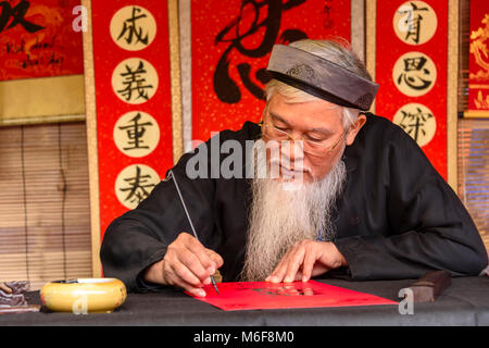 An elderly Vietnamese man with a long white beard uses a brush to draw Chinese characters calligraphy in Hanoi, Vietnam Stock Photo