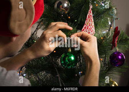 Young male with masquerade funny hat on his head decorates christmas tree for celebrating  family holiday Stock Photo