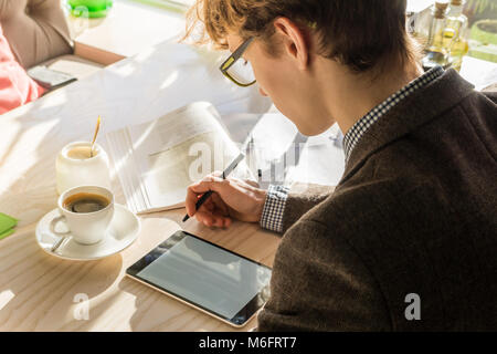 Doing home assignment project at cafe during lunch. Young modern male teenage student works with tablet pc and books on bright sunny day in cozy restr Stock Photo