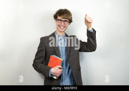 Smiling young male student makes thumbs up gesture. Handsome teenage person in tweed jacket and eyeglasses holds school books and laughs in sincere em Stock Photo