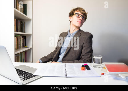 Handsome young person in reading eyeglasses at modern minimalistic workplace rests and turns away from table, looking up. Young male student works wit Stock Photo
