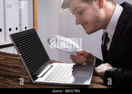 Young Businessman Looking At Binary Code With Magnifying Glass On Wooden Desk Stock Photo