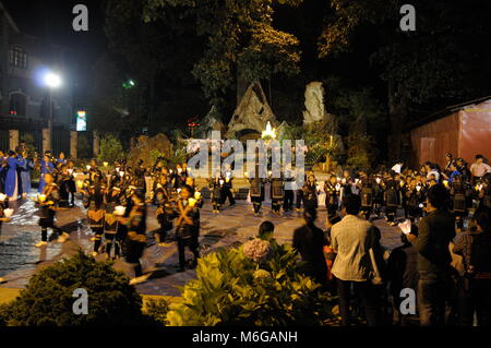 Young happy and solemn Hmong girls dressed in traditional clothing holding candles and flowers celebrating Virgin Mary at Sapa Stone Church at night. Stock Photo