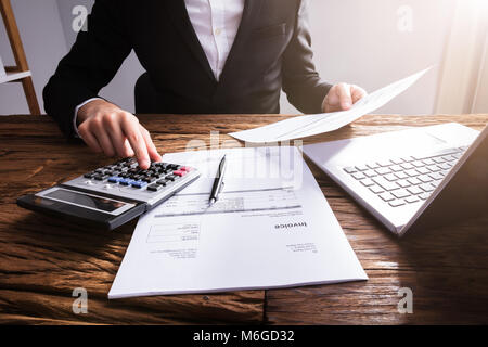 Businessperson's Hand Calculating Invoice With Laptop On Wooden Desk Stock Photo