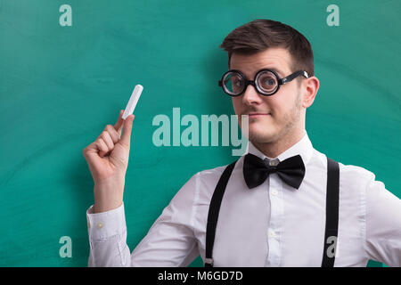 Close-up Of A Young Male Teacher Holding Chalk Against Green Chalkboard Stock Photo