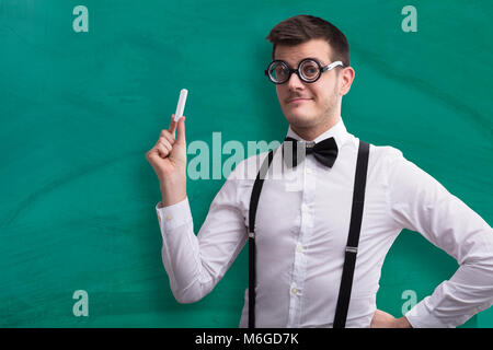 Close-up Of A Young Male Teacher Holding Chalk Against Green Chalkboard Stock Photo