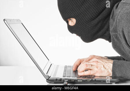 Male hacker in balaclava writing virus code on laptop keyboard Stock Photo