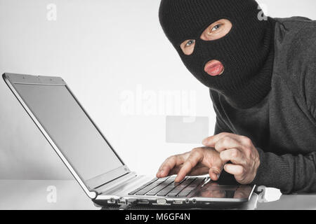 Male hacker in balaclava holding a card in left hand and typing something with right hand on laptop keyboard Stock Photo