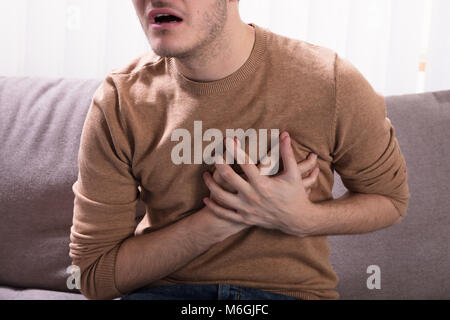 Close-up Of A Young Man Sitting On Sofa Having Heart Attack  At Home Stock Photo
