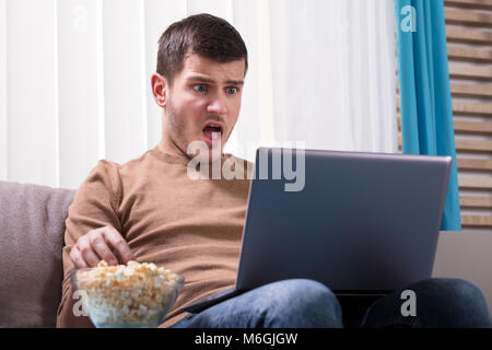 Shocked Man Looking At Laptop And Eating Popcorn At Home Stock Photo
