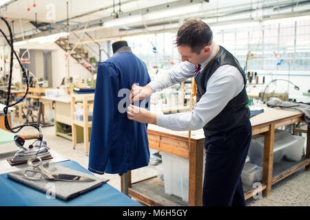 Tailor working on a partially completed new blue jacket on a mannequin in his large spacious workshop tucking in the pleats at the back with his hands Stock Photo