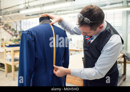 Tailor measuring the length of the back of a new blue jacket on a mannequin that he is busy sewing in his workshop using a tape measure Stock Photo