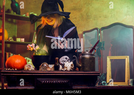 Portrait of witch in black hat reading spell over pot with green steam Stock Photo