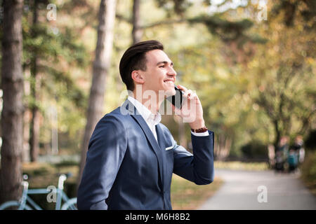 Young businessman talking on smartphone outdoors in a park, happy and smiling. Stock Photo