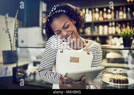 Smiling young African entrepreneur standing at the counter of her cafe talking on a cellphone and using a tablet Stock Photo