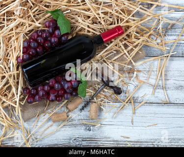 Overhead view of a red wine bottle, grapes plus corkscrew with straw and burlap on white rustic boards Stock Photo