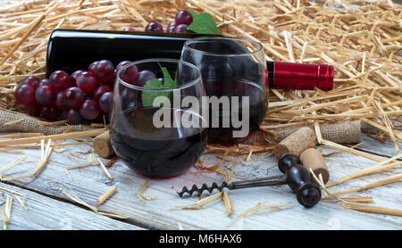 Close up view of drinking glasses filled with red wine, bottle and grapes plus corkscrew with straw and burlap on white rustic boards Stock Photo