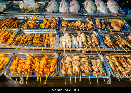 Fried chicken meat on skewers and fresh fish are offered to eat in illuminated street restaurants at night in the centre of town Stock Photo
