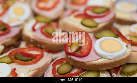 Open-face sandwiches with sausage and vegetables as background. Close-up of white bread slices with ham, cheese, eggs, capsicum and cucumber. Stock Photo