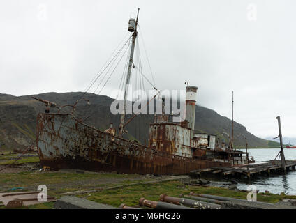 An old, rusted whaling boat no longer in use on the shore at Grytviken, South Georgia. Fog, a hillside and the bay are in the background. Stock Photo