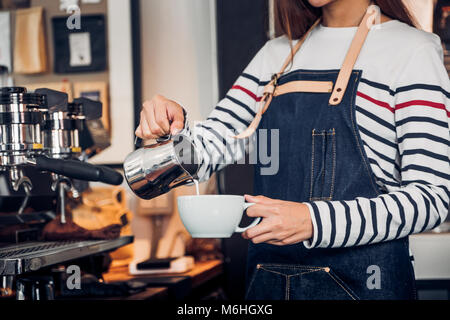 Asia woman barista pour milk into hot coffee cup at counter bar in front of machine in cafe restaurant,Food business owner concept Stock Photo