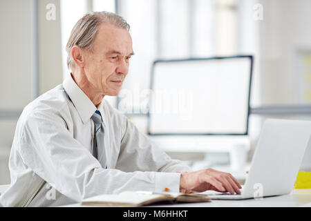 Authentic elderly office worke typing on laptop in office with glass walls Stock Photo