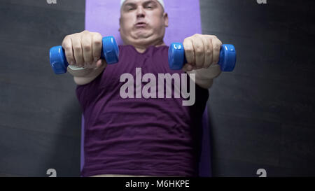 Top view of overweight man lying on floor and lifting dumbbells with effort Stock Photo
