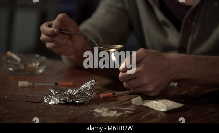 Addict heating heroin dose in spoon with lighter, injectors and white lie around Stock Photo