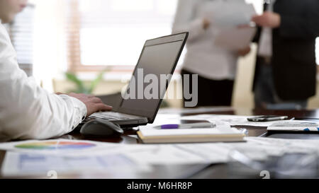 Worker taking notes on laptop while two colleagues discussing papers at office Stock Photo