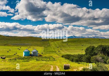 Small silos, bales of hay at ranch in rolling foothills of the Rocky Mountains, visible in far distance, near Longview, Alberta, Canada Stock Photo