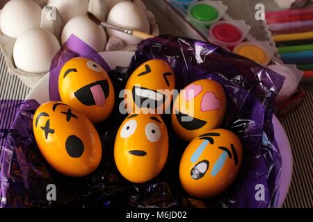 Emoticons Easter Eggs on purple cellophane paper. More white eggs, brush and paints in the background. Stock Photo