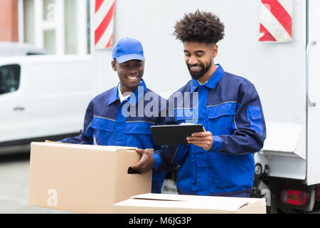 Two Delivery Men Standing Near The Van Looking At The Digital Tablet With Cardboard Boxes Stock Photo