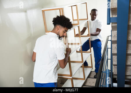 Two Male Movers Carrying The Empty White Shelf On The Staircase At Home Stock Photo