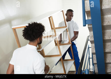 Two Male Movers Carrying The Empty White Shelf On The Staircase At Home Stock Photo