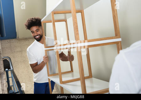 Two Male Movers Carrying The Empty White Shelf On The Staircase At Home Stock Photo