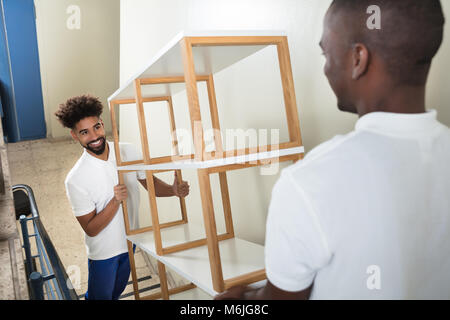 Two Male Movers Carrying The Empty White Shelf On The Staircase At Home Stock Photo