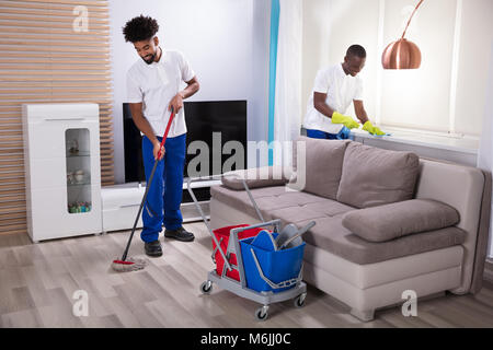 Two Smiling Young Male Janitor Cleaning The Windowsill And Mopping Floor In The Living Room Stock Photo
