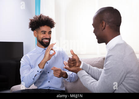 Two Young Happy Men Sitting On The Sofa Making Sign Languages Stock Photo