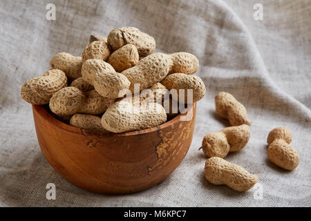 Composition of raw unpeeled peanuts in clay ceramic bowl on light grey cotton tablecloth or napkin, some copy space, closeup, selective focus. Healthy Stock Photo