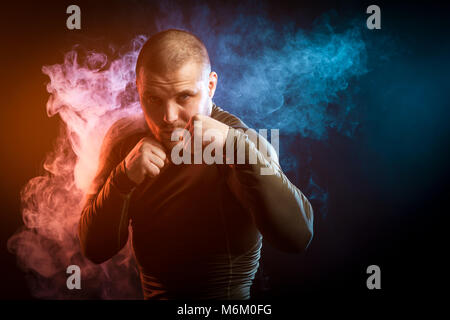 A strong young dark-haired male athlete in a green sports jacket is boxing against a background of blue and red smoke on a black isolated background Stock Photo