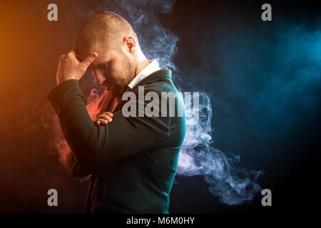 Young handsome businessman man in white shirt and green jacket thought over problem solving opposite red and blue smoke from wipe on black isolated ba Stock Photo